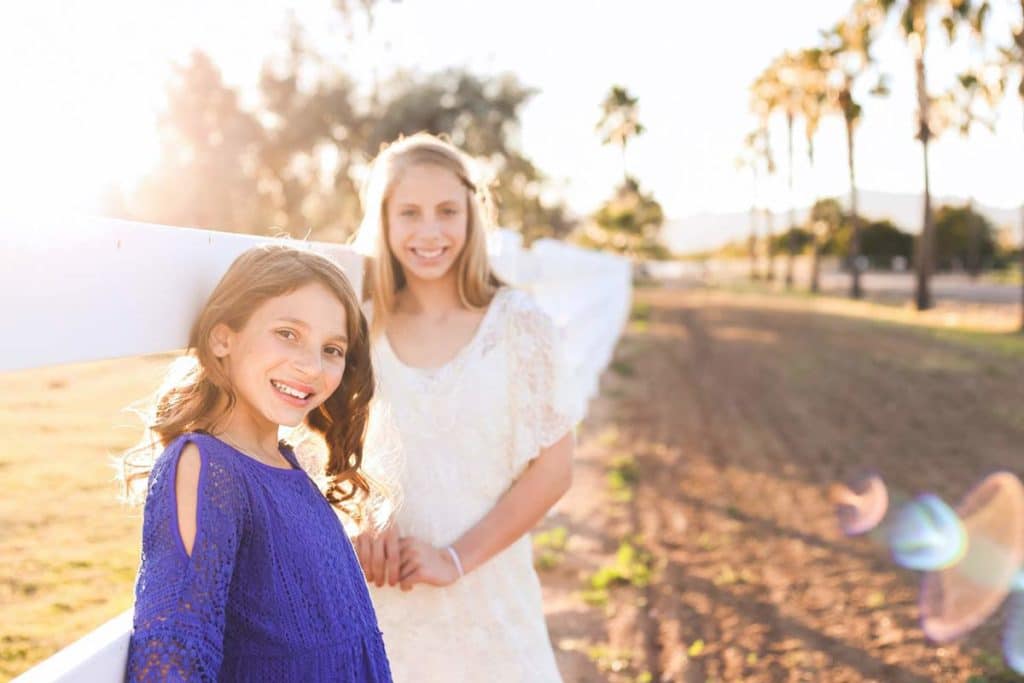 gorgeous photograph of two sisters blue and white dresses outside at family farm with sun shining setting by white picket fence for sibling moment captured by north mississippi photographer danielle jacqueline