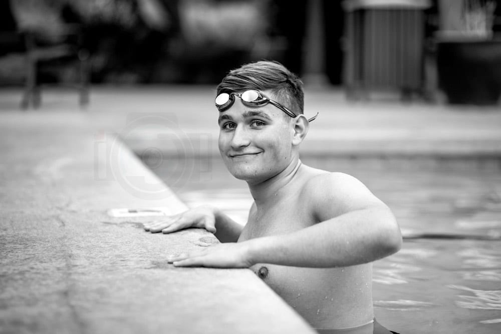 angle from left looking down pool deck at high school swimmer hands on edge smiling with goggles on head for graduation photo session with danielle jacqueline near memphis tennessee