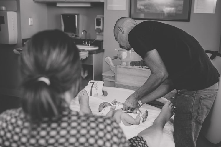 first time dad changing newborn baby daughter's diaper in hospital room with mother looking on black and white photo child crying candid by danielle jacqueline photography desoto county mississippi