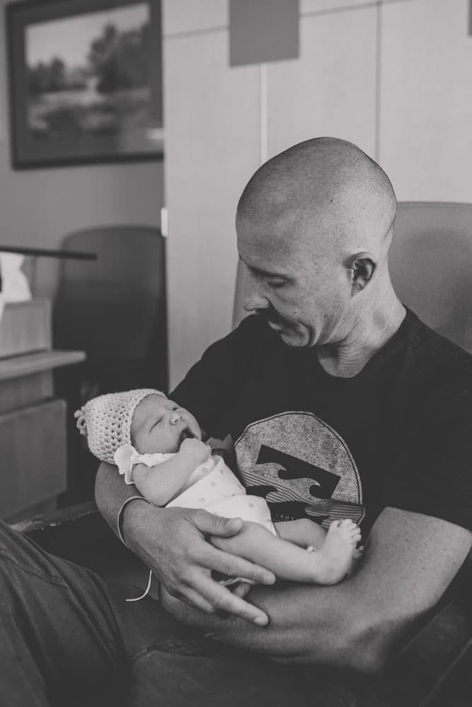 intimate moment of daddy with first daughter in hospital chair post delivery room black and white staring at baby's eyes enjoying moment captured by photographer in olive branch mississippi