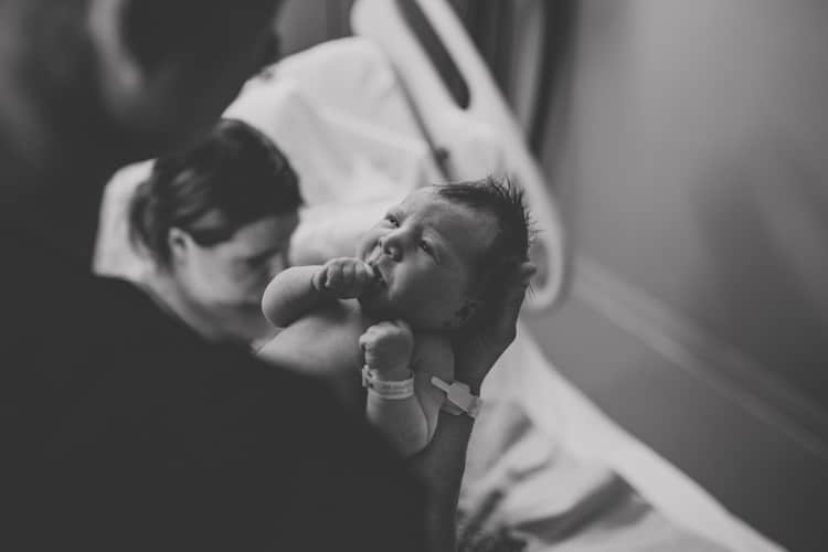 close up of newly born baby girl in father's arms looking into his eyes while mother prepares to breastfeed on hospital bed by olive branch mississippi photographer danielle jacqueline black and white