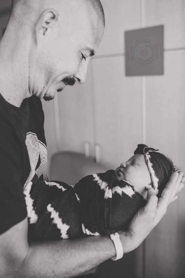 folder delicately holding newborn baby daughter from side arms bent looking down black and white at hospital with photographer danielle jacqueline near memphis tn