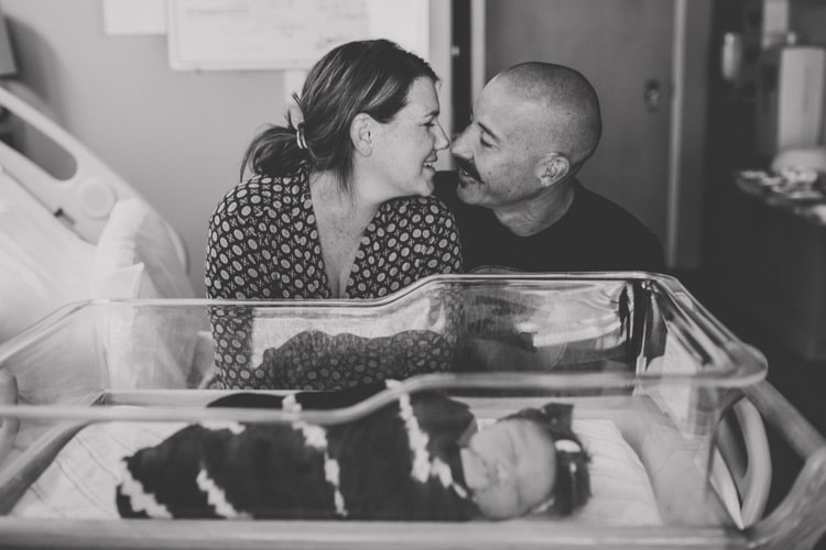 loving parents going in for kiss black and white photo with newborn baby in incubator in front of them at hospital for intimate moments of parents in olive branch mississippi by danielle jacqueline