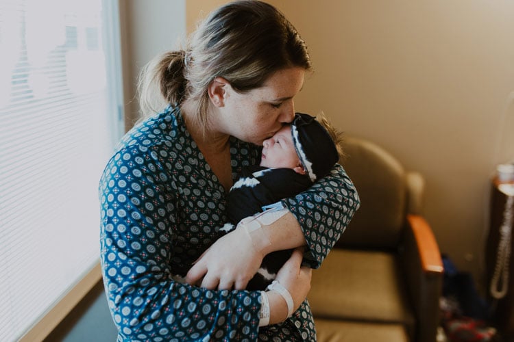 mom kissing first baby on forehead by hospital room window during professional photoshoot with infant photographer danielle jacqueline in olive branch mississippi