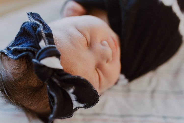 close up portrait of infant girl in delivery room after birth for photography session with parents blue bow tied on head swaddled tight sleeping mississippi