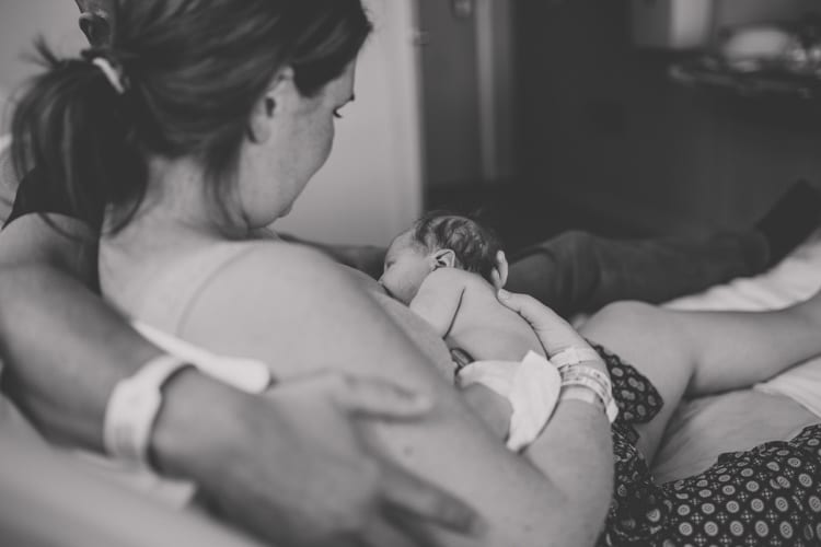 over the shoulder over first time mom as she breast feeds her new born baby daughter in hospital with husband's arm around her in hospital bed intimate black and white photography near memphis tn by danielle jacqueline