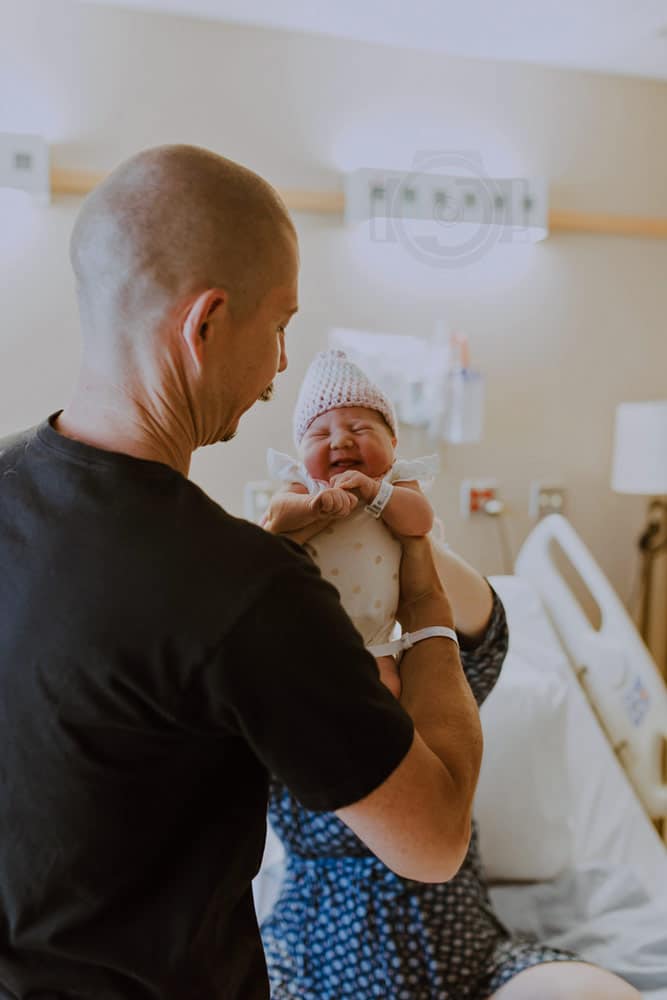 first time father taking baby hand off from mother who is laying in hospital bed during fresh 48 session with danielle jacqueline photography located in desoto county ms