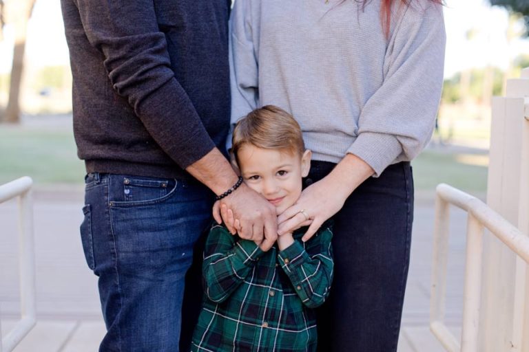 waist down of parents eye level with 5 year old boy in between both of them holding onto their hands outdoors by white railing wearing green and blue flannel for holiday greeting card photography by olive branch mississippi based danielle jacqueline