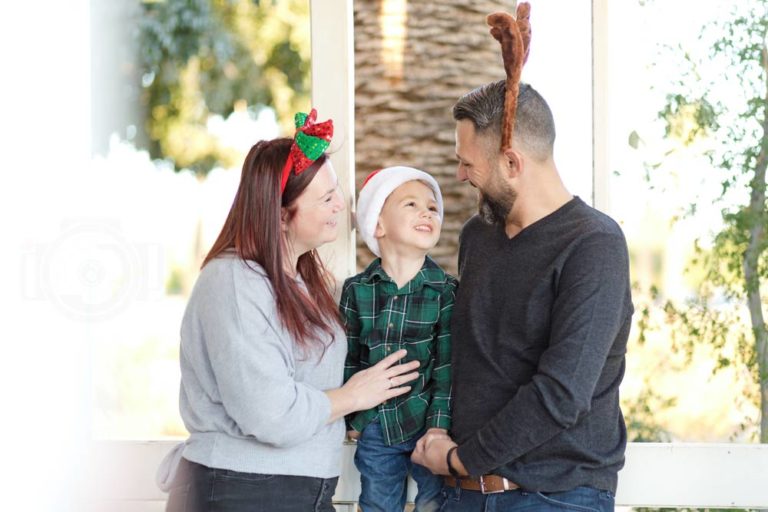 son looking up into father's eyes while sitting on white fence next to large tree while mom watches them both wearing christmas props on heads for family holiday photography session by danielle jacqueline in olive branch ms