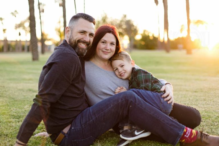 daddy seated on grass with mom in front of him and son on her lap as they look right towards camera with sun setting beyond trees in background creating golden hue for family holiday pictures with danielle jacqueline photography in desoto county ms