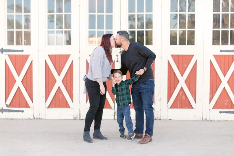 mom and dad kissing over the top of 5 year old only son as he holds their hands in front of brand new red and white barn doors for family holiday pictures by desoto county photographer danielle jacqueline mississippi