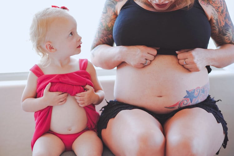 mommy to be in olive branch mississippi sitting down with 2 year old daughter during maternity portrait session wearing pink and black window in background by danielle jacqueline photography