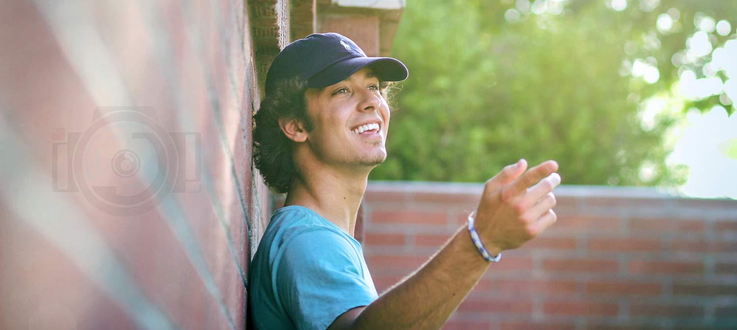 senior photo session in olive branch ms with teen in ball cap pointing off to right leaning against brick wall with perfect lighting and bright green tree behind him by danielle jacqueline photography