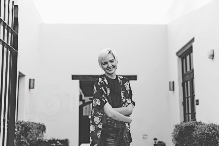 high school senior walking out of house in flower shawl laughing while looking off to the light arms crossed courtyard for black and white senior portrait by danielle jacqueline lifestyle out of olive branch mississippi