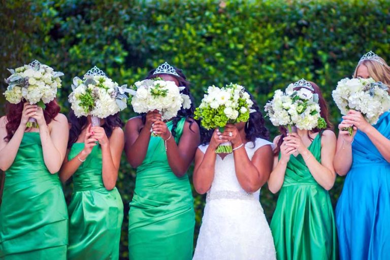 bride with bridesmaids holding beautiful boquets of flowers over their faces during wedding photographer session in memphis tennessee wearing green and blue dresses bright green shrubs in background at popular resort by danielle jacqueline