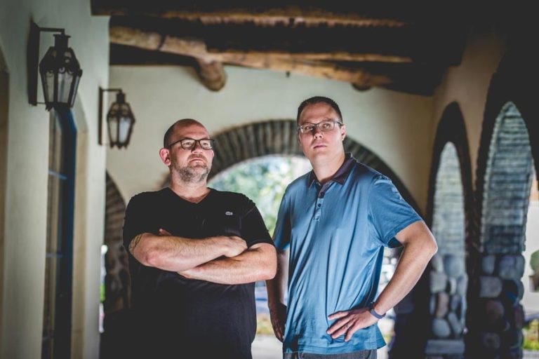 two entrepreneurs standing under arched outdoor walkway of office building for business olive branch photography services by danielle jacqueline wearing blue polo hands on hips and blank t shirt arms crossed successful podcast hosts nationwide professionals