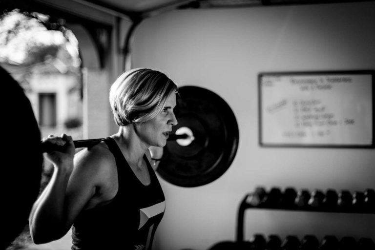 black and white image of business owner standing at entrance of gym overhead doors squat rack determined look on face with workout regimen on white board next to dumbbell rack photography bu danielle jacqueline in olive branch ms