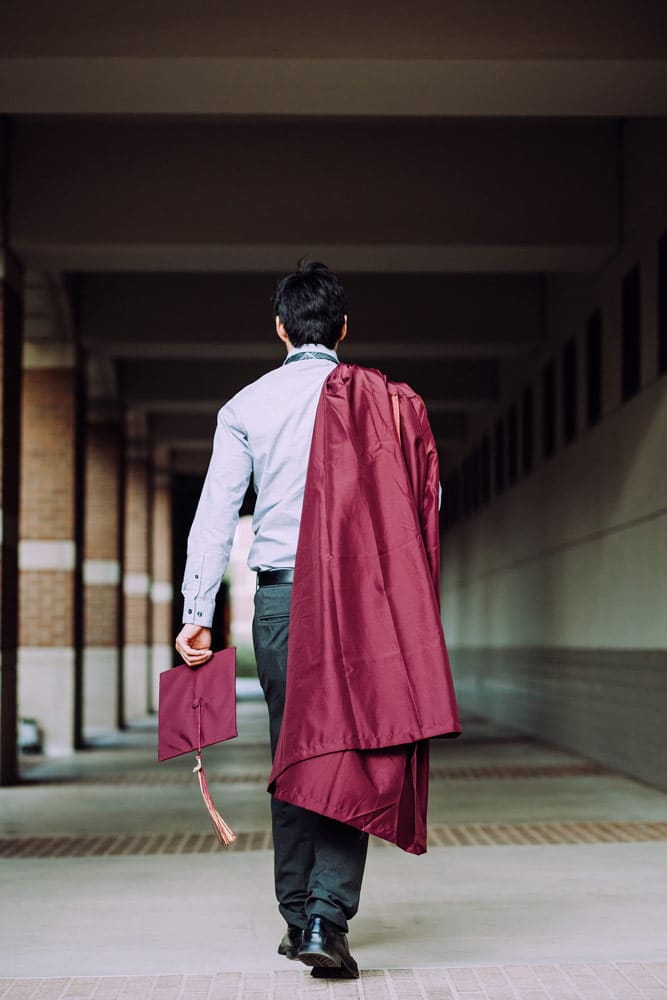 college graduate walking away from desoto county photographer while carrying diploma and gown over right shoulder and tassel on cap in left formal attire