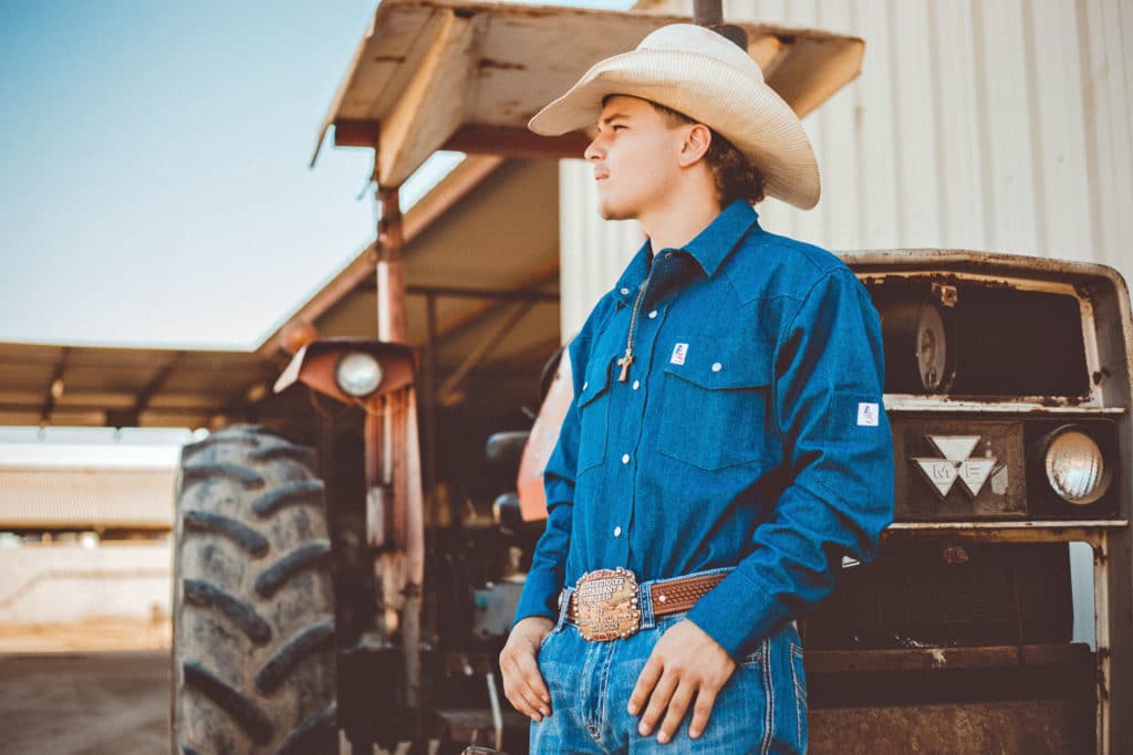 thumbs in belt loops belt buckle cowboy hat denim long sleeve in jeans looking away left leaning against tractor for apparel business photography session by danielle jacqueline in olive branch ms