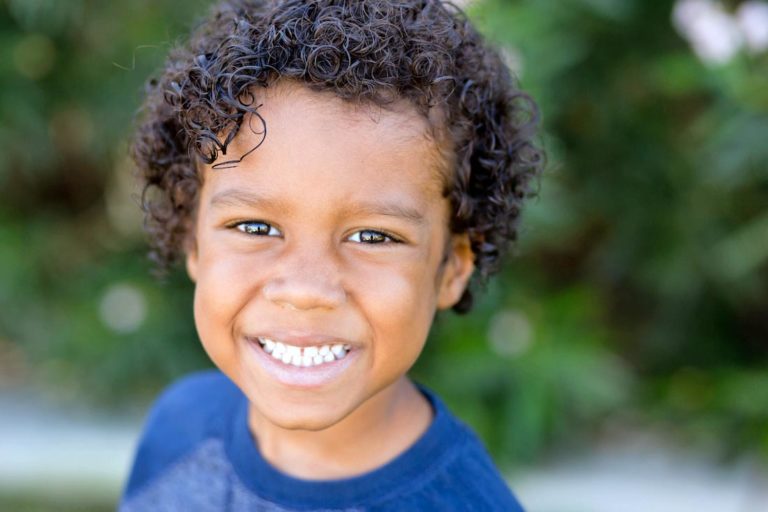 curly perm hair young boy smiling big for back to school photos with danielle jacqueline children's photographer trees in background blue shirt in germantown tn