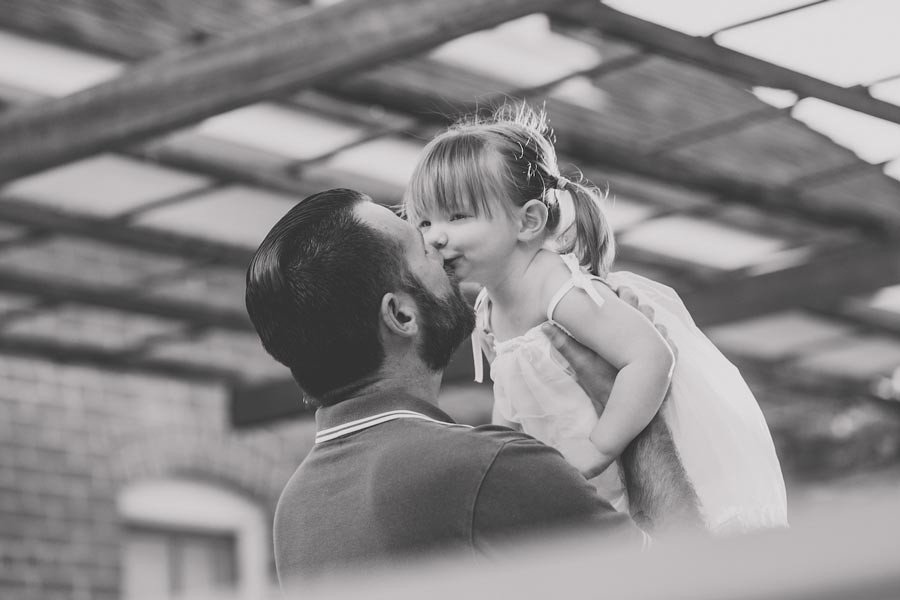 loving daddy holding up baby girl in his arms trellice showing in ceiling of historical park candid moment kissing pig tailed white dress great angle capturing fence blur by danielle jacqueline photography family portraits desoto county ms