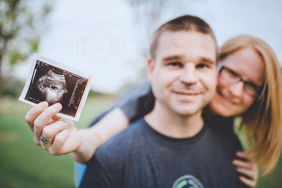 wife holding ultrasound image from doctor's visit to announce having a baby with her husband in olive branch mississippi portraits done by danielle jacqueline photography at park with grassy background and blue skies