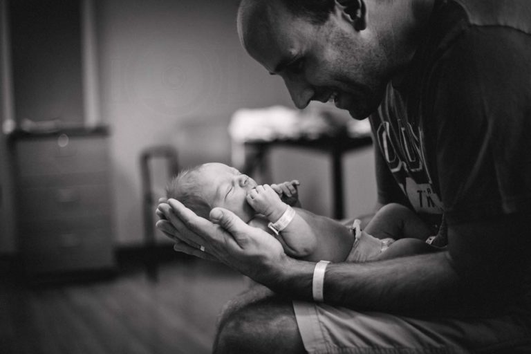 happy daddy palms up holding and looking at newly born son at hospital with physician equipment in background in awe of God's creation captured by danielle jacqueline photography in memphis tn