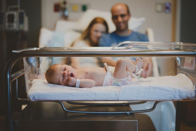 new mother and father sitting on hospital bed looking at baby arrival in clear tub at feet in diaper cooing together as married couple after hiring danielle jacqueline photography for fresh 48 session in memphis tn