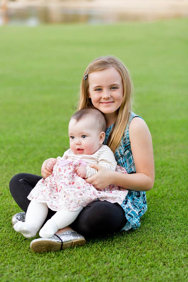 happy big sister with new baby sister on lap sitting in grass dressed up for family session with danielle jacqueline photography in southaven ms