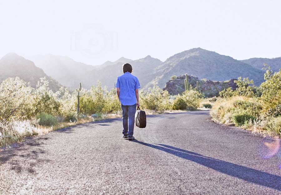 high school senior boy photos walking down winding road in hills no traffic with suitcase aspiring musician blue shirt jeans glasses no worries for proud parents keepsake by graduation photographer in desoto county ms