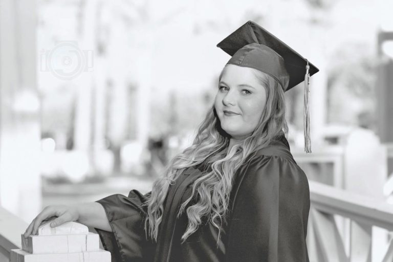 cap and gown blank stare to reflect on accomplishments of young woman black and white senior photos of mississippi graduates by danielle jacqueline hand on bridge railing