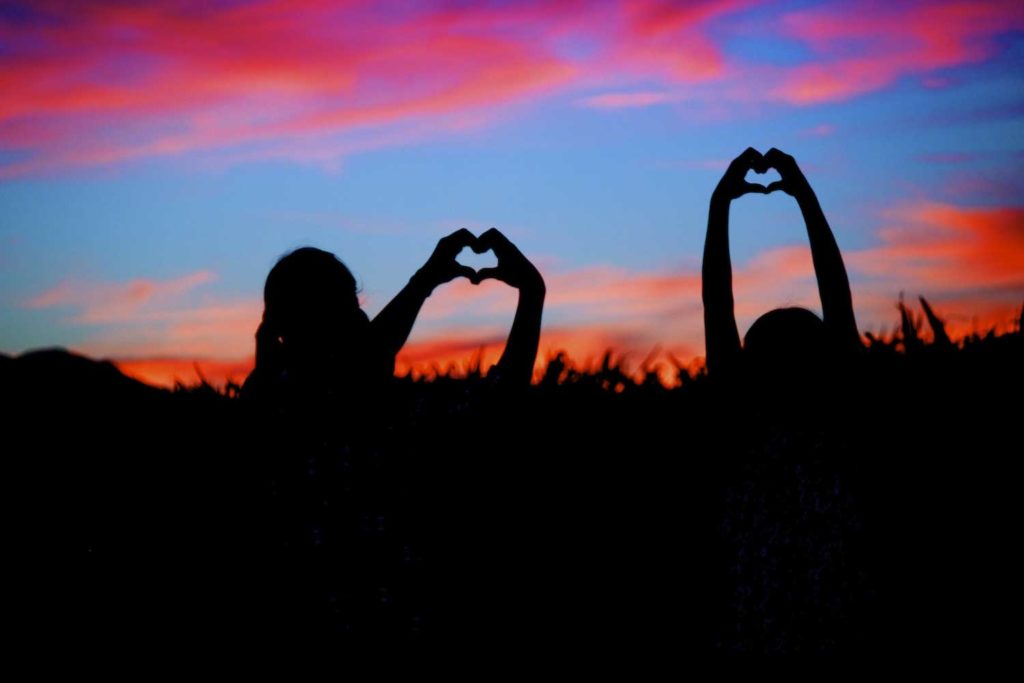 daughters of olive branch photographer in ms holding hands up to create heart shaped silhouette in front of cornfield blue sky with purple pink and orange clouds at dusk