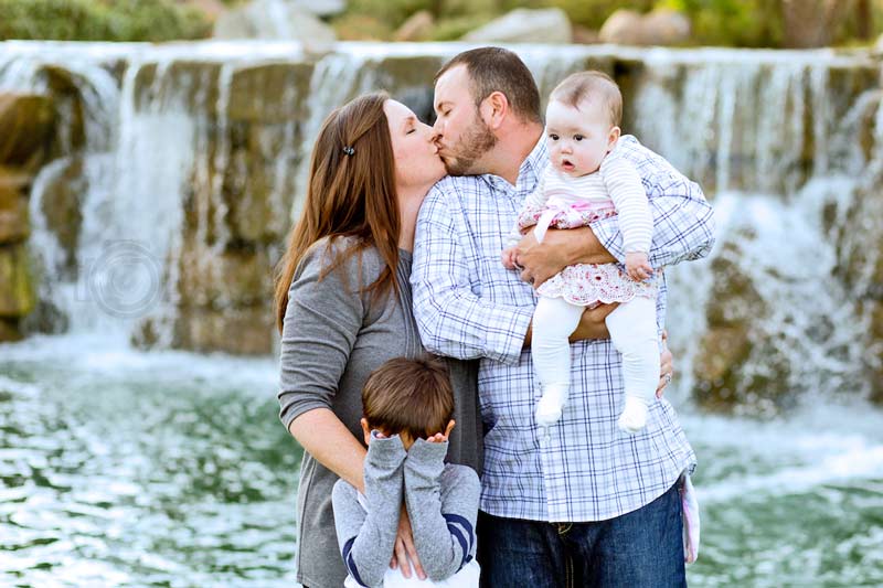 mom and dad giving each other a kiss in front of water fall in memphis tn neighborhood while son hides eyes and baby daughter looks annoyed for family photos by danielle jacqueline