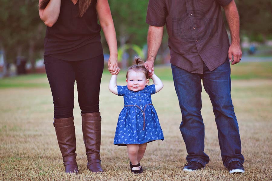 mommy and daddy in maroon walking with 1 year old daughter in grass at park during the fall for family newborn pictures by danielle jacqueline photography outdoors