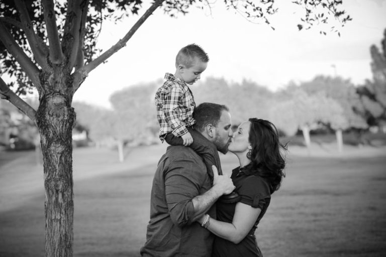 little boy on shoulders of father at park by tree black and white as he kisses his wife for personal family portraits by danielle jacqueline photography in desoto county ms