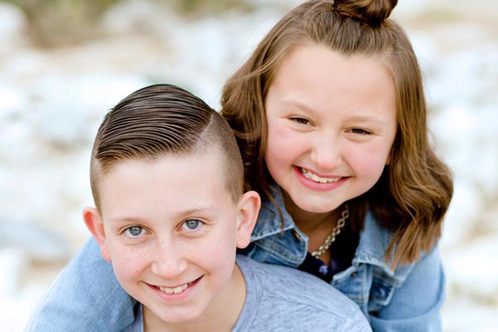 sister behind brother looking up at camera with stack of big rocks in background during outdoor kids photographer session with danielle jacqueline in horn lake ms