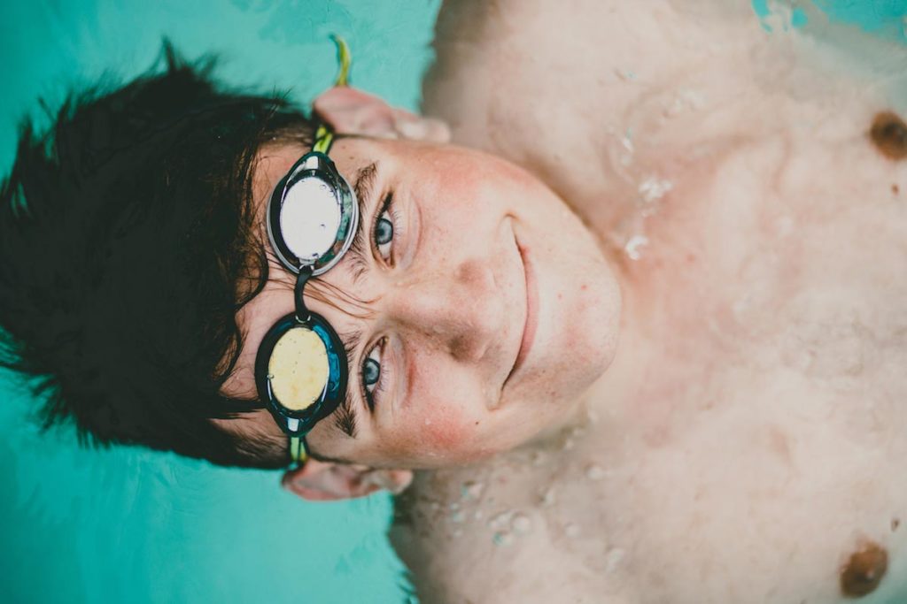 looking up at camera in water with goggles over eyes big grin hair floating for swim team senior portraits in olive branch ms by danielle jacqueline for boys creative student athletes