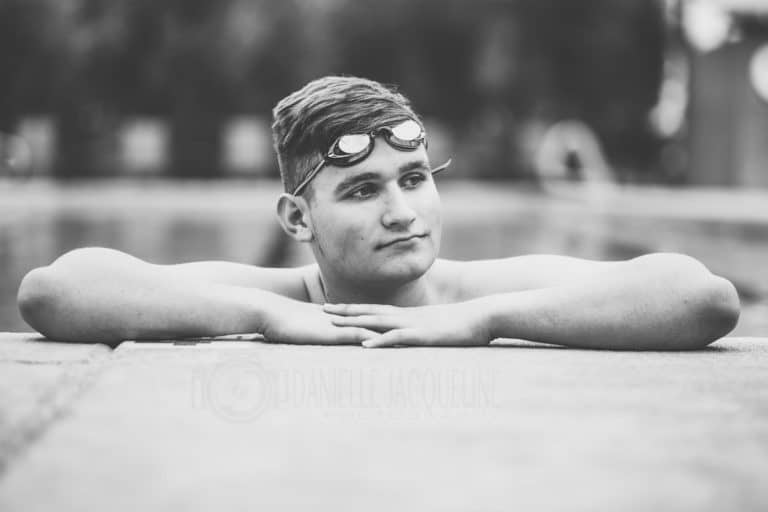 background image for danielle jacqueline photography services of high school senior posing in swimming pool with arms on ledge and goggles on before graduation student athlete looking right for parents in olive branch mississippi