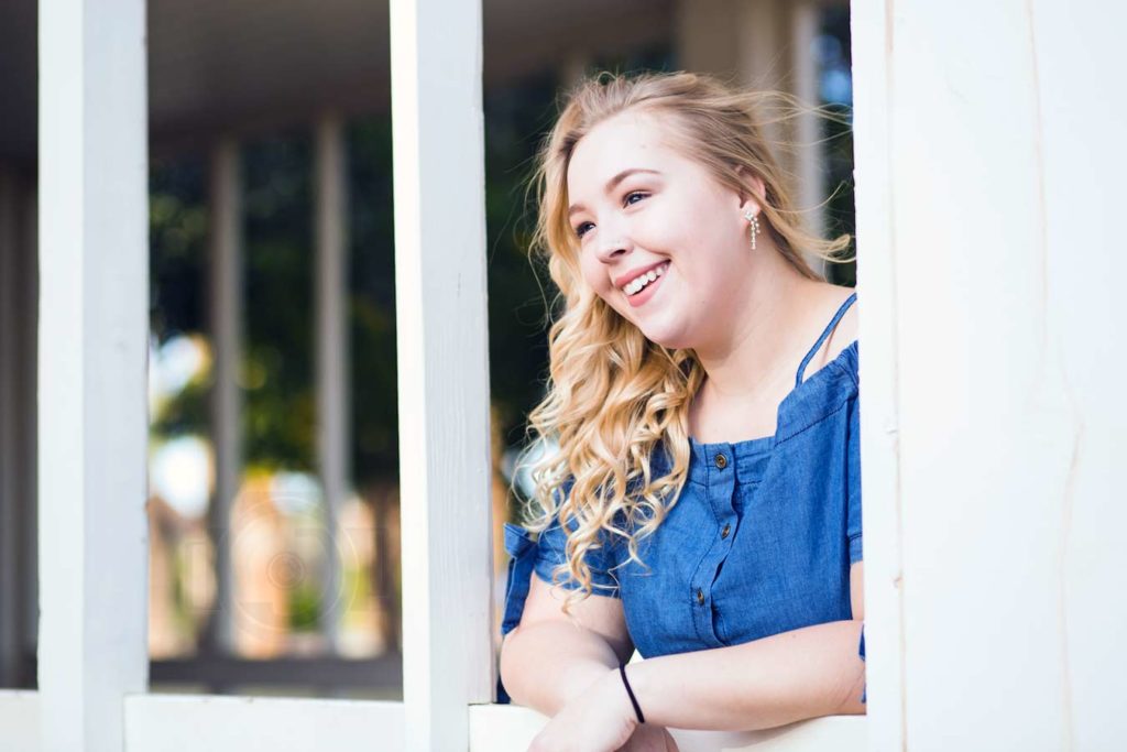 farmgirl senior photo session in denim dress hair done leaning on porch painted white for graduation celebration with olive branch ms photographer danielle jacqueline