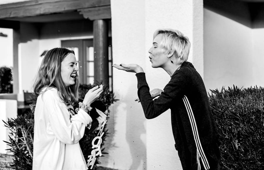 adult short haired blonde daughter blowing glitter off hand into face of mom laughing outside of house during special event for family black and white image by danielle jacqueline