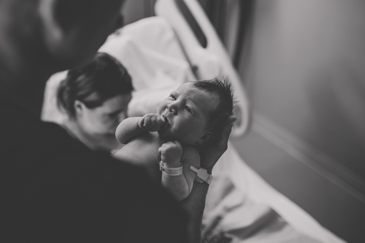 black and white newly born baby looking up at father while he hold daughter and birthing mother in background on hospital bed captured by danielle jacqueline photography