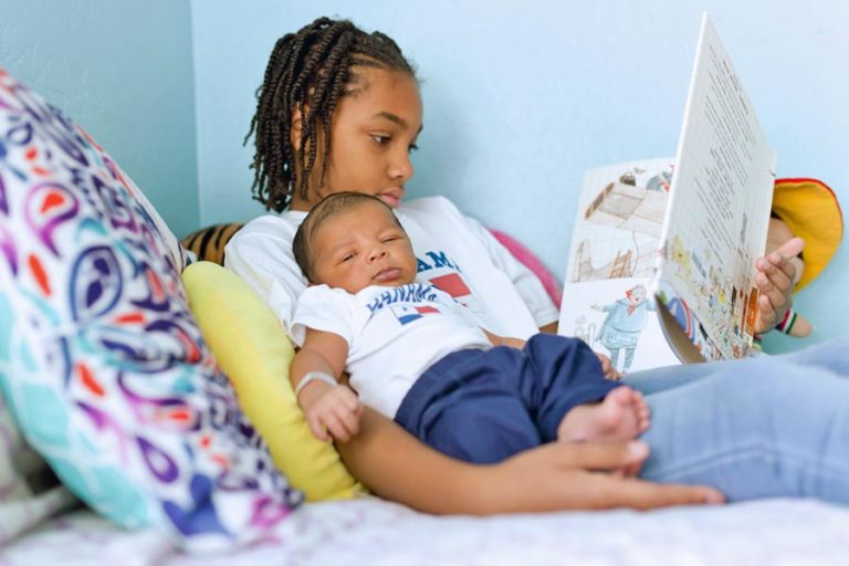 first time big sister reading her little newborn brother a story on her bed during in home photoshoot with danielle jacqueline