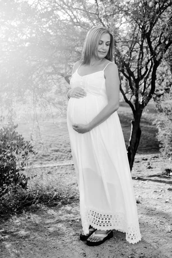 black and white portrait of pregnant mother in sun dress in front of trees with sun light flair resting on her like halo outdoor by danielle jacqueline photography in desoto county ms