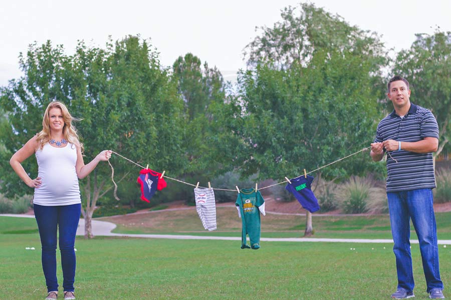 holding up clothesline of baby boy clothes during maternity photography session outside with husband and wife couple expecting son in desoto county green grass trees hands on hip smiling mom