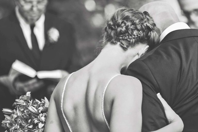 bride to be clinching her father's arm as he prepares to give his daughter to marriage at alter with pastor reading scripture and gorgeous hairstyle from behind by wedding photographer in olive branch ms danielle jacqueline