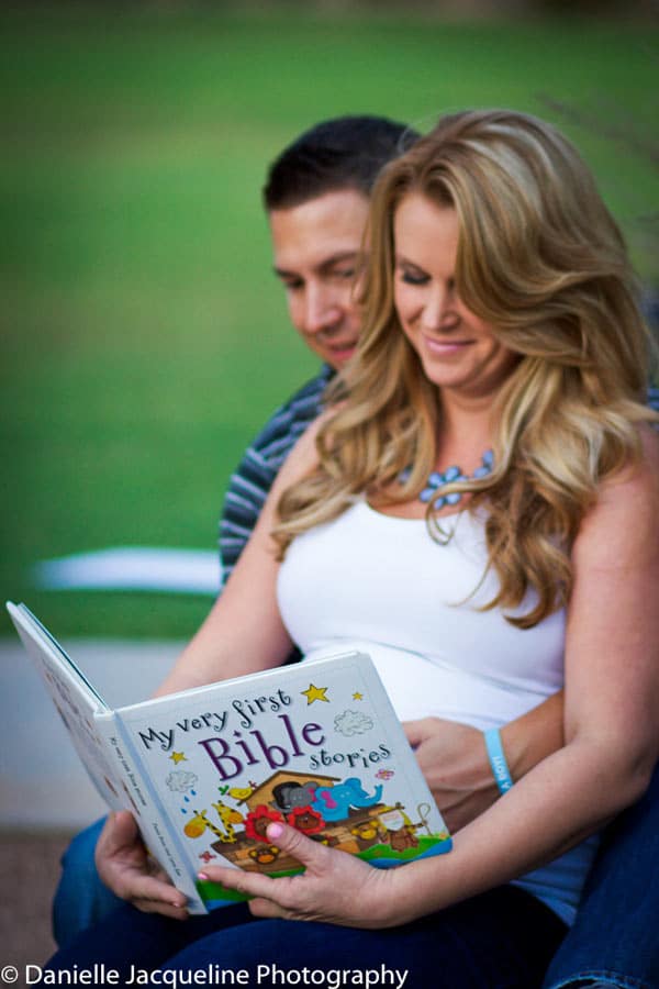 wife expecting child sitting on husbands lap reading God's word outside on park bench for maternity portraits by danielle jacqueline photography green grass in background natural lighting hair done