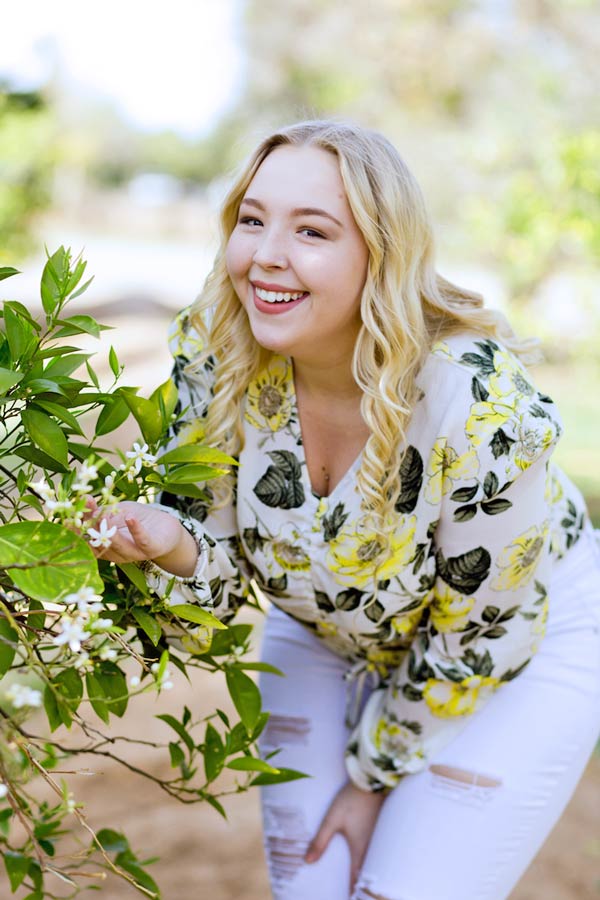 smelling the rose pedals at family orchard in northern mississippi wearing white pants flowered green and yellow blouse with bright blonde hair for head shot modeling auditions by danielle jacqueline