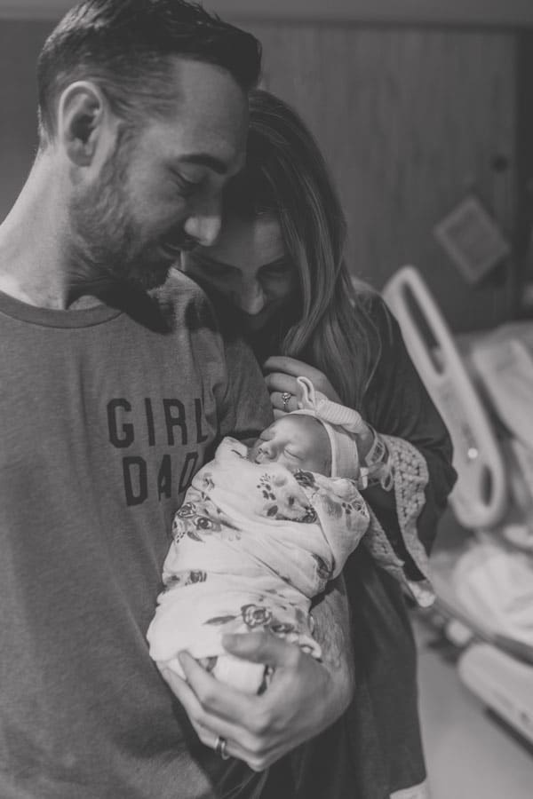 mommy and daddy leaning against each other foreheads standing next to hospital bed looking down in awe at new baby girl arrival in their arms by desoto county ms black and white photographer danielle jacqueline