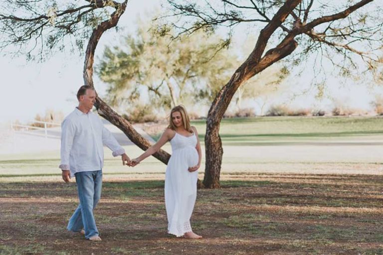 walking away from husband under trees as he's holding her hand dressed in all white expecting father in blue jeans for maternity portraits in southaven ms by danielle jacqueline