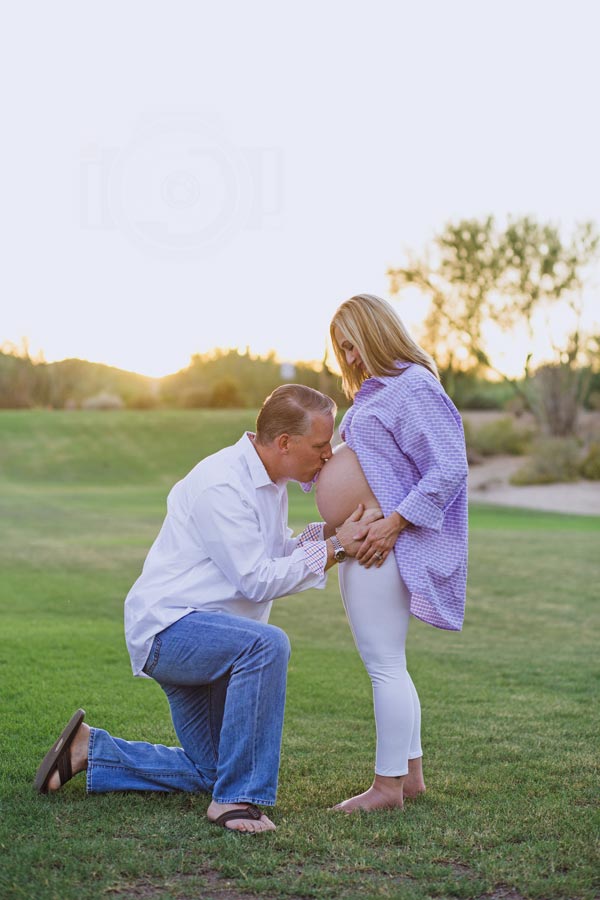 husband middle age on knee in jeans and white shirt in open grassy park kissing wife's belly for maternity portrait session outdoors color by danielle jacqueline photography in olive branch ms